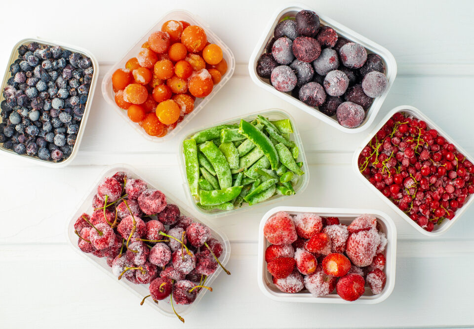 Frozen berries and vegetables in plastic boxes on white wooden background.